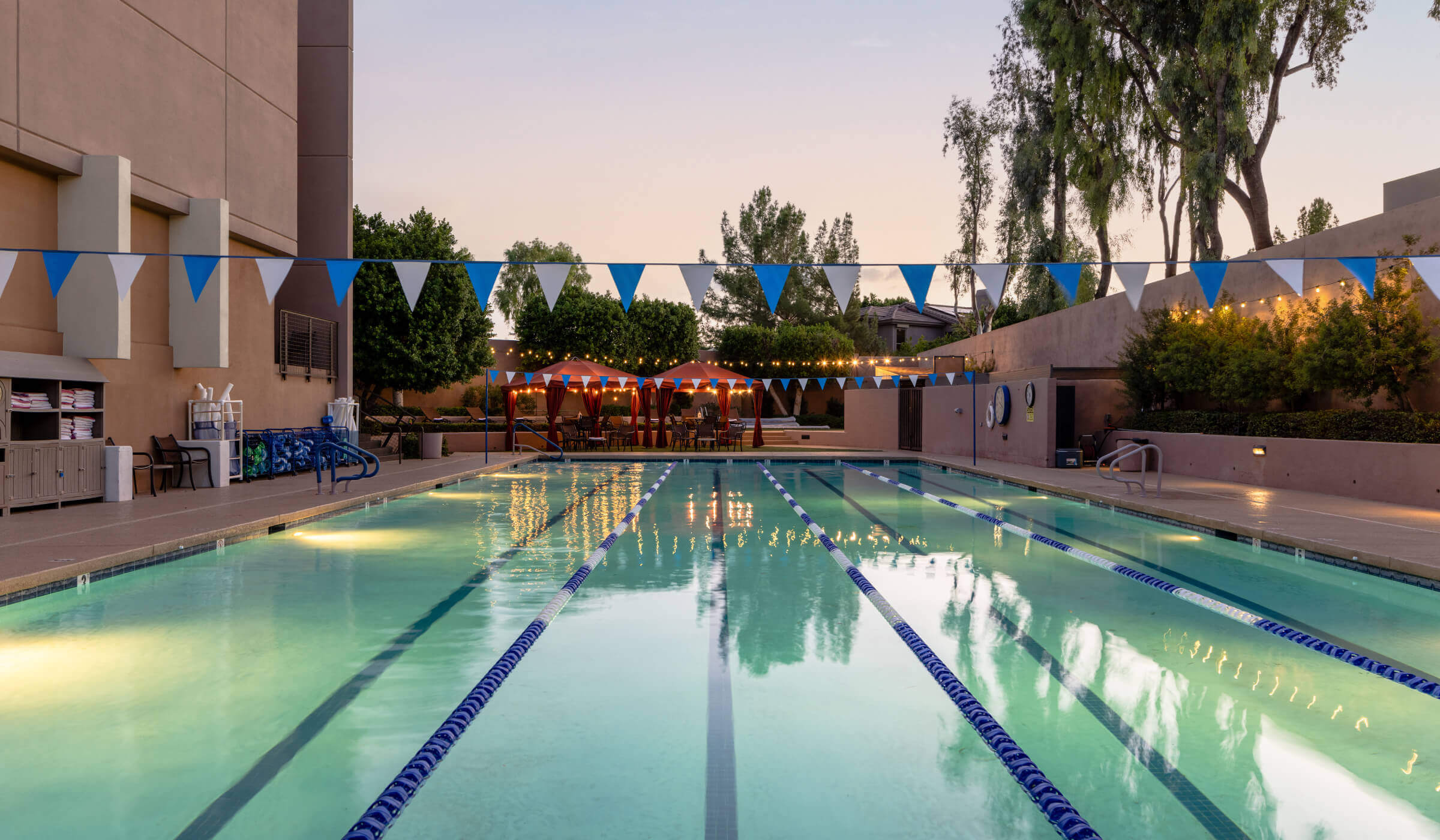Village Clubs Camelback outdoor lap pool at dusk, with lane markers, blue-white flags, trees, and lounge chairs in view.