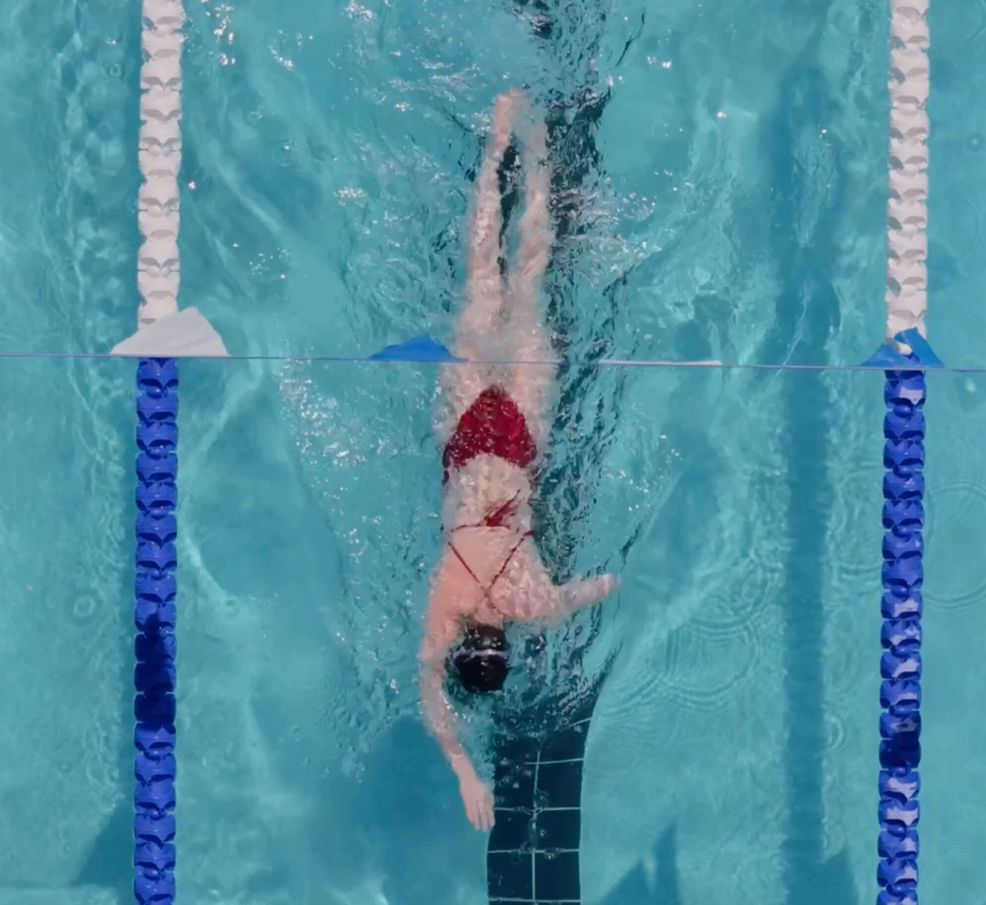 A swimmer in a red suit swims freestyle at Village Clubs Camelback’s clear blue pool, between lane dividers, viewed from above.