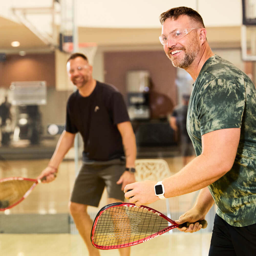 Two men in safety glasses play racquetball at Village Clubs Camelback’s indoor court, smiling and holding racquets.