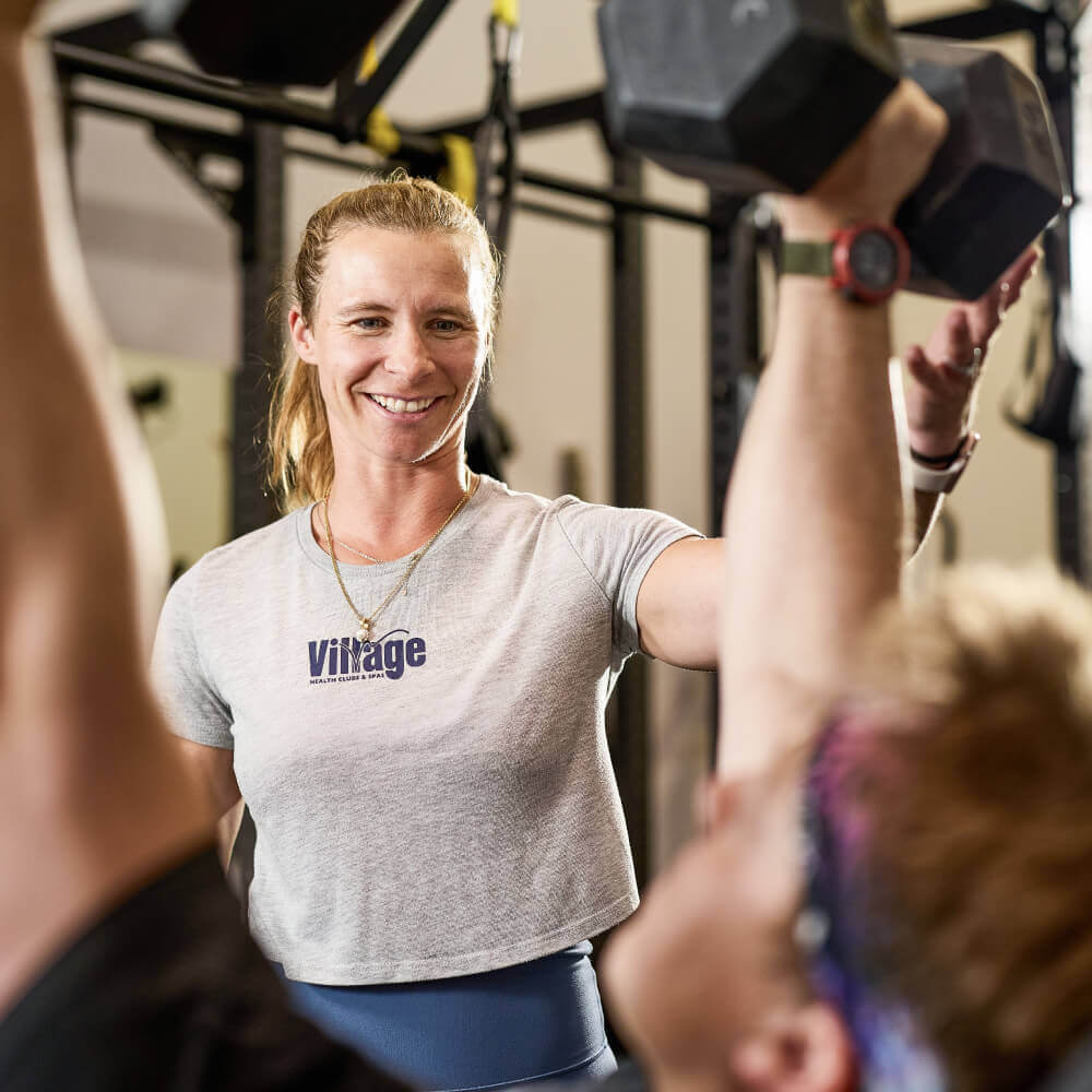 A Village Clubs trainer at Camelback smiles while coaching a client lifting dumbbells in a gym with equipment visible behind them.