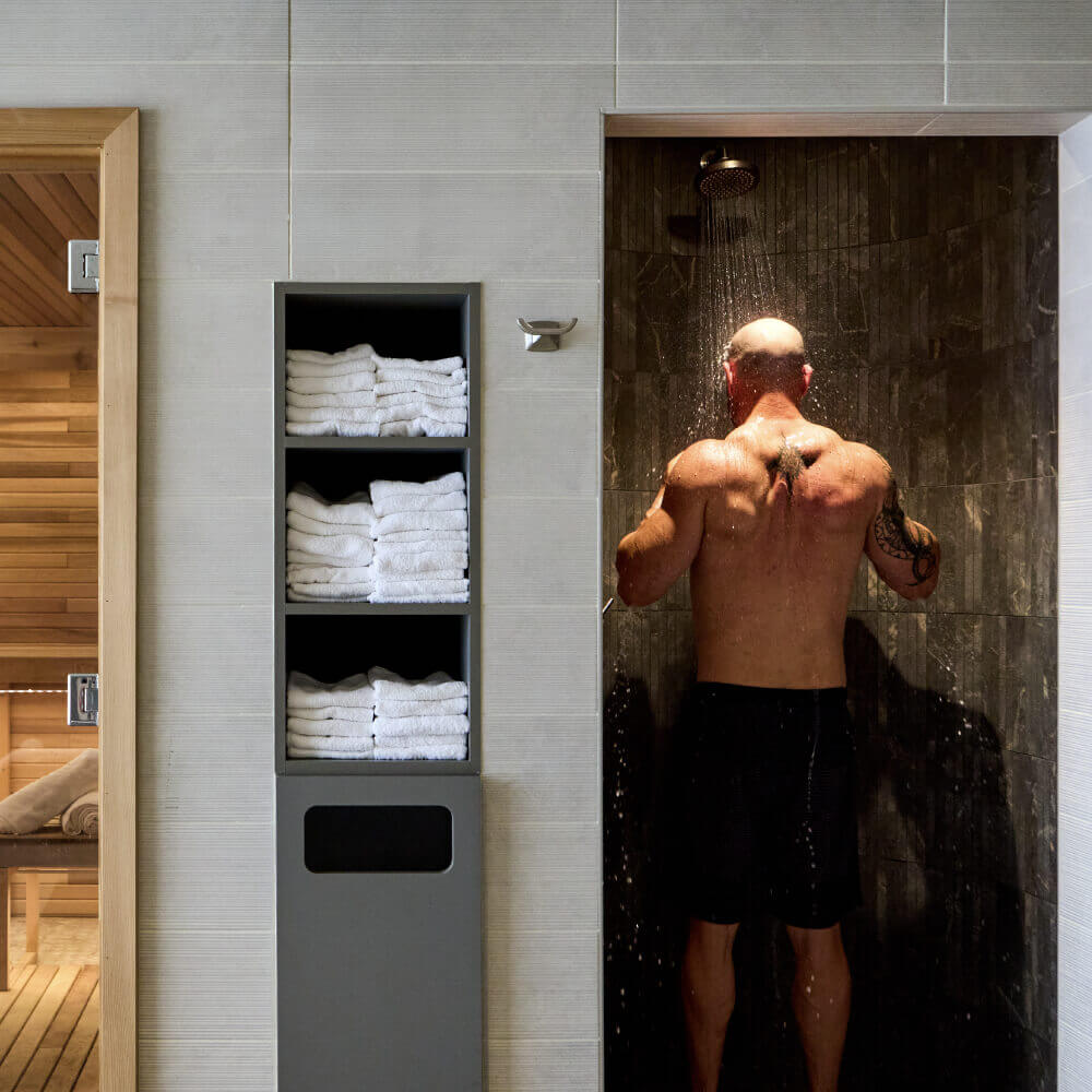 A man showers at Village Clubs Camelback, with dark tiles, white towels on shelves, and a sauna room visible nearby.