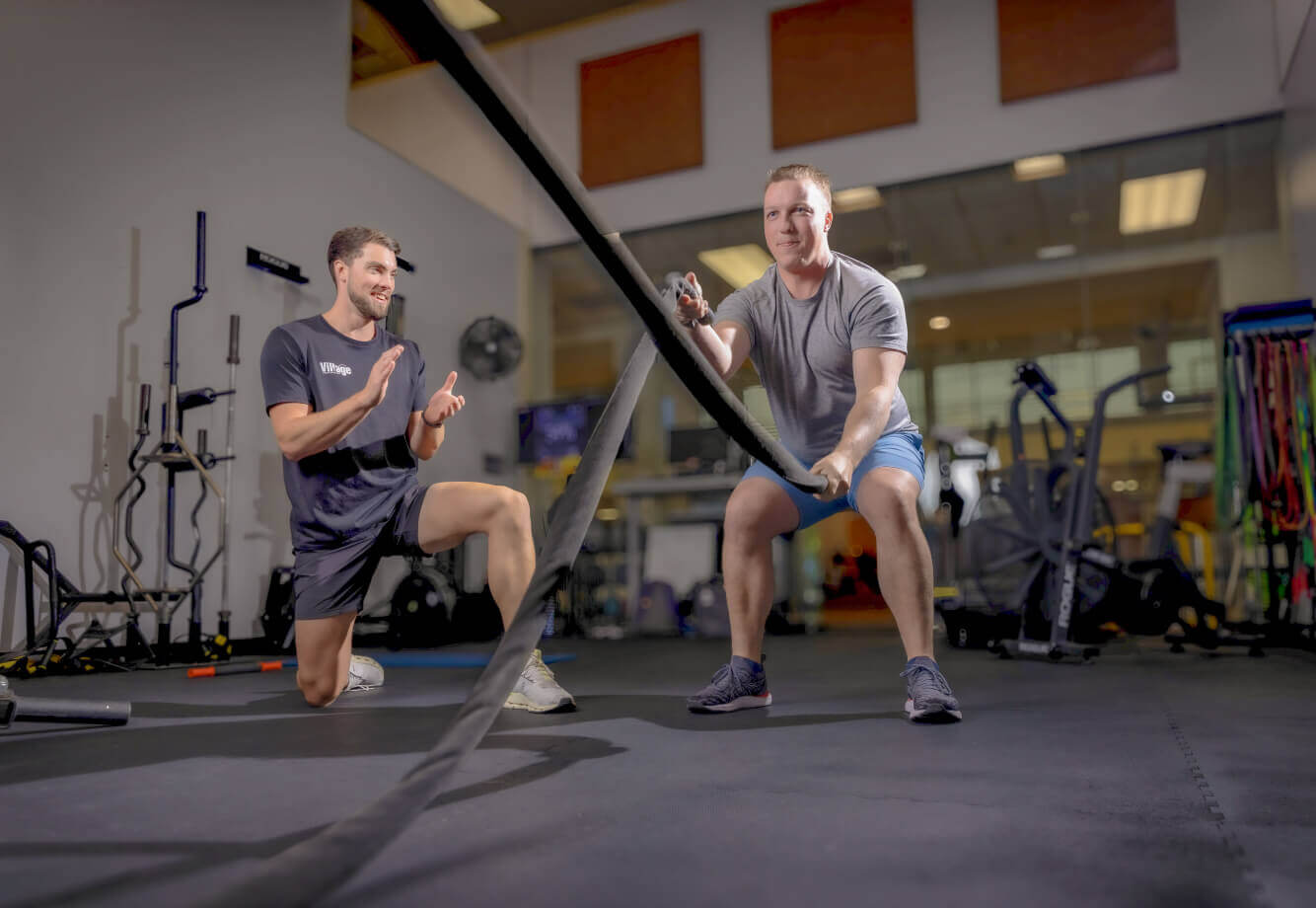 At Village Clubs Camelback, a coach kneels and smiles while training a man using battle ropes in the gym’s fitness area.