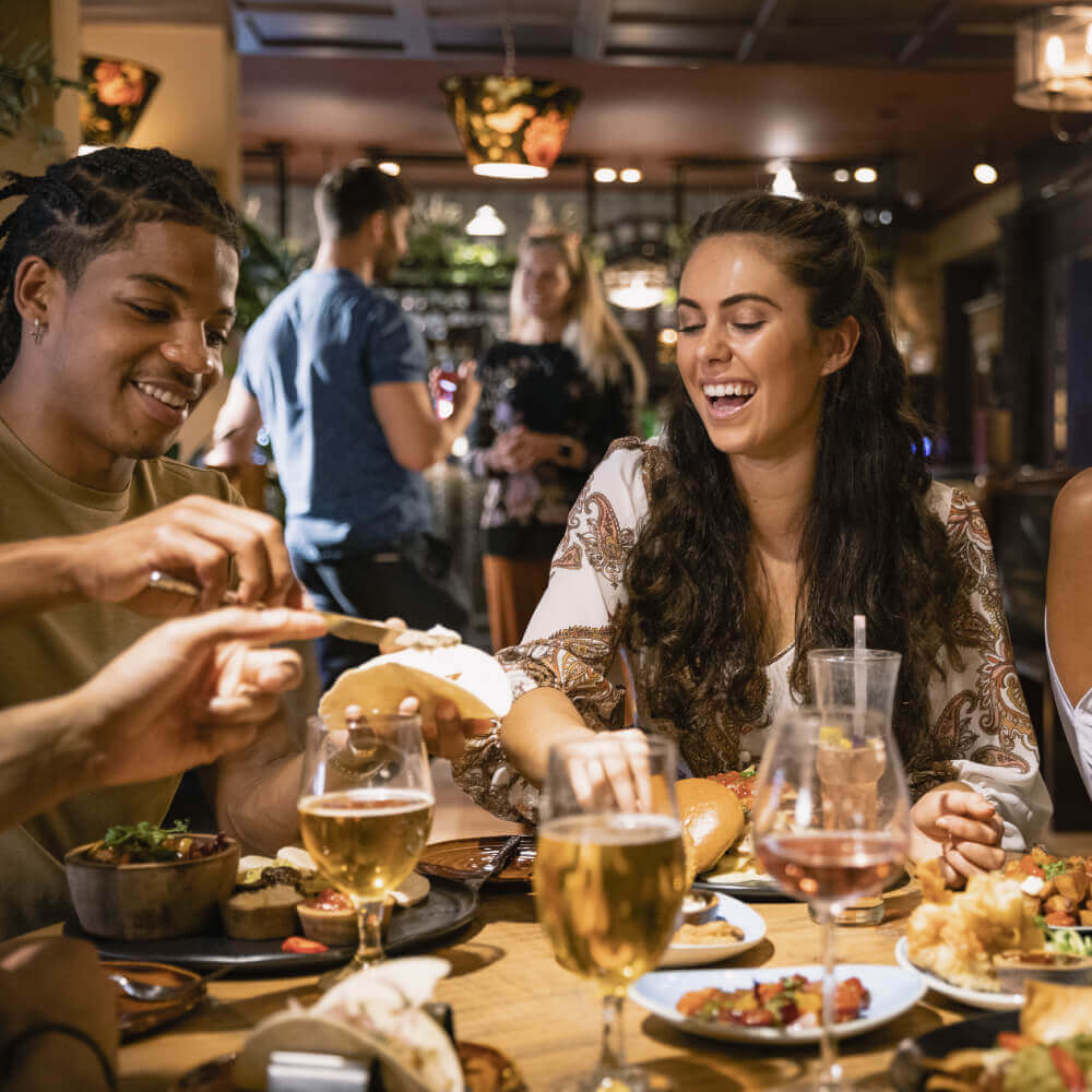 Friends enjoy dining and laughter at Village Clubs Camelback, with drinks, food, and a lively, warmly lit restaurant atmosphere.