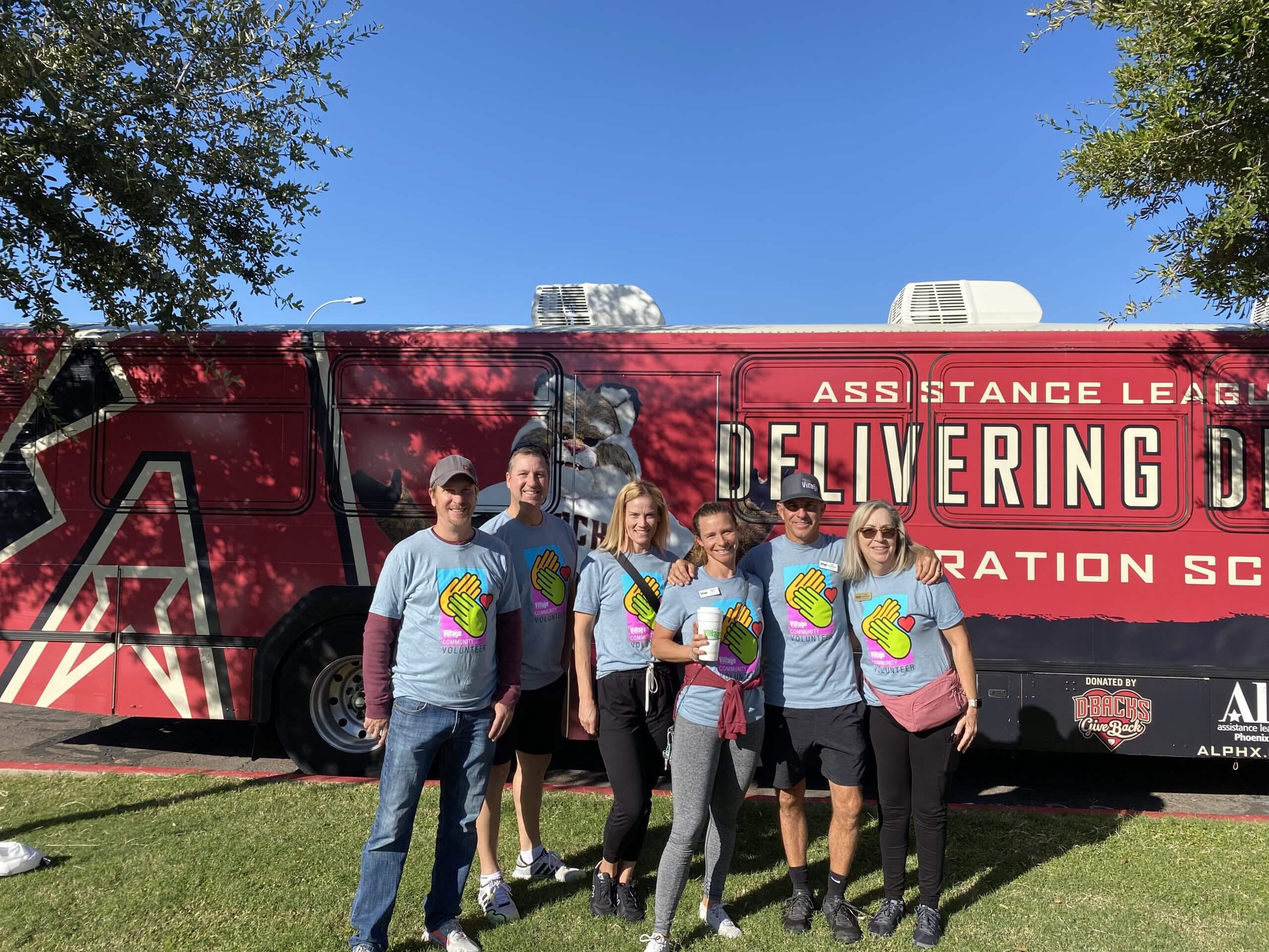 Six Village Clubs Camelback team members in blue Village Cares shirts smile by an Assistance League bus on a sunny day.