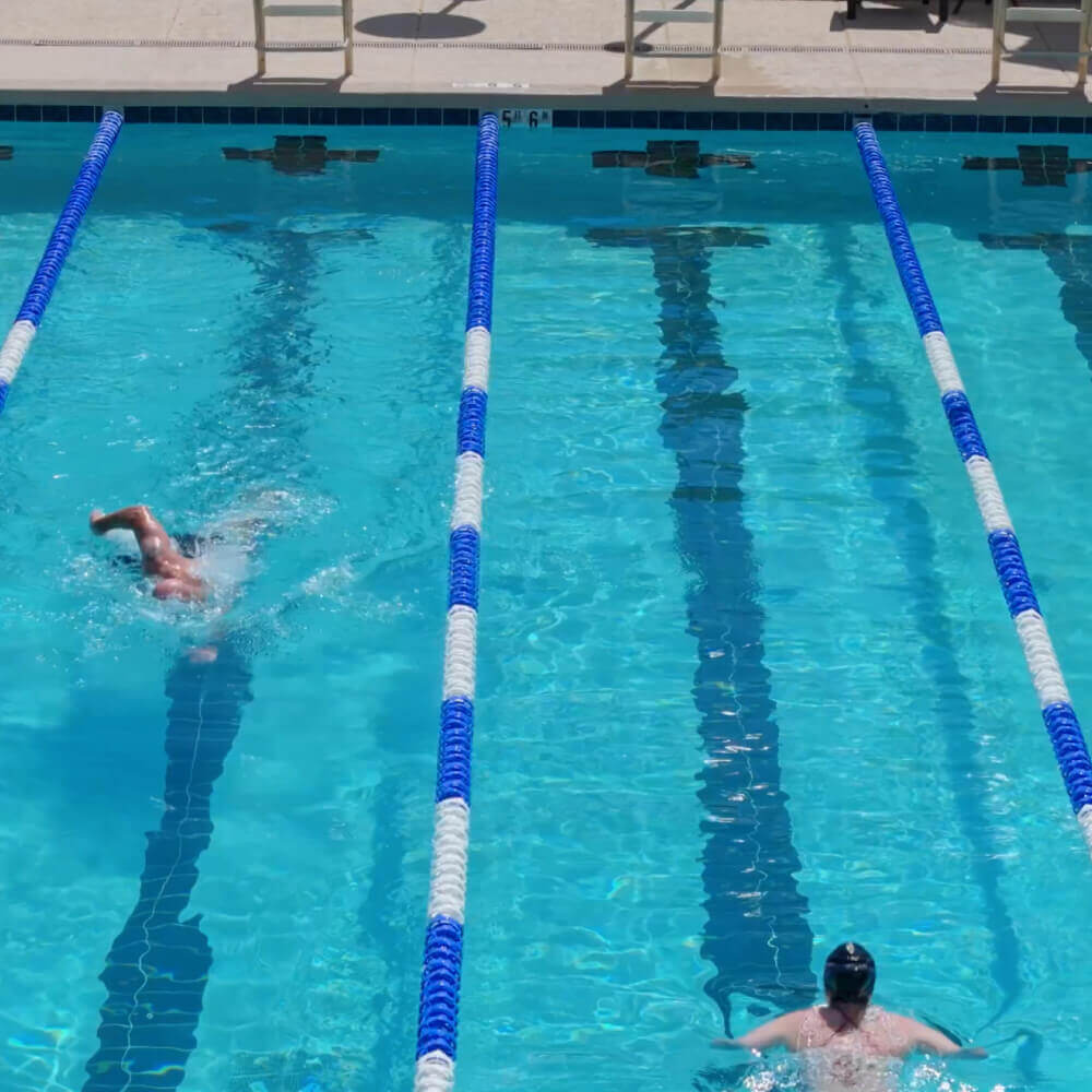Swimmers do laps in the clear outdoor pool at Village Clubs Camelback, with lane dividers, chairs, and deck in view.