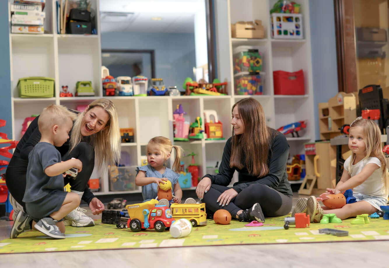 At Village Clubs Camelback, two women and three children play together on a colorful mat surrounded by toys in the playroom.