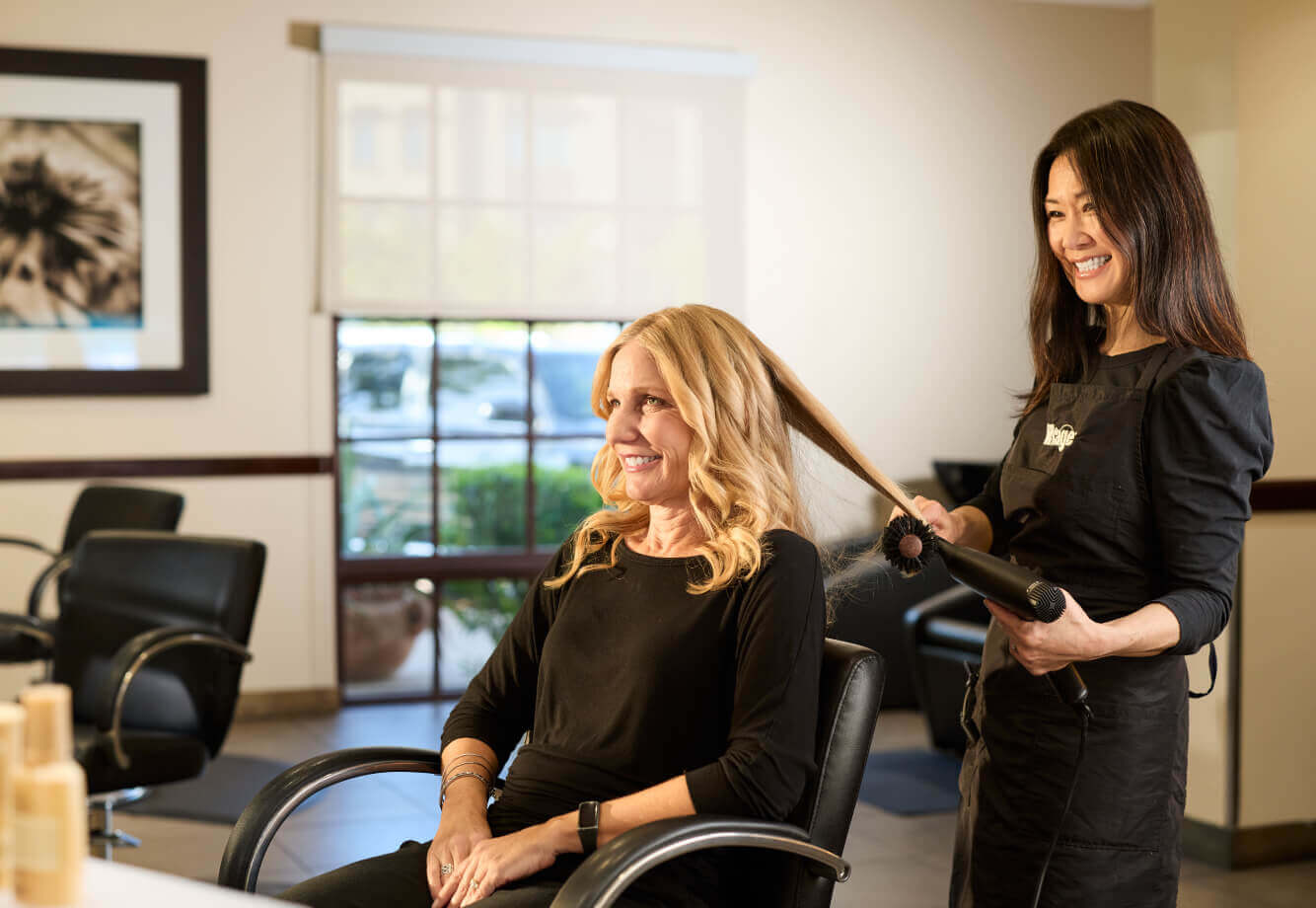 A stylist at Village Clubs Camelback curls a guest’s blonde hair in a bright, modern salon; both women are smiling and relaxed.
