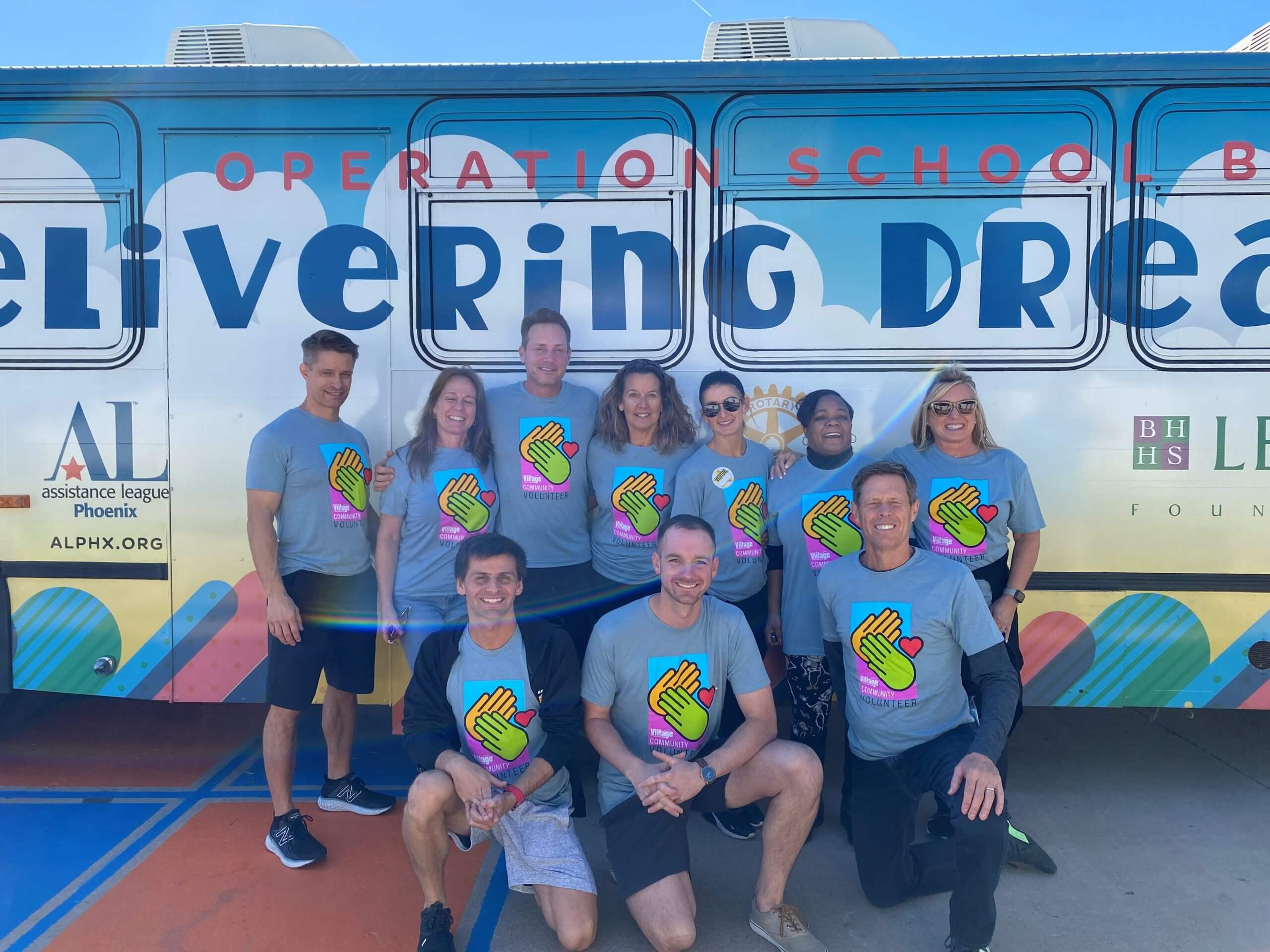 Ten smiling people in Village Cares shirts pose by a colorful bus at Village Clubs Camelback, with logos and a bright blue sky.