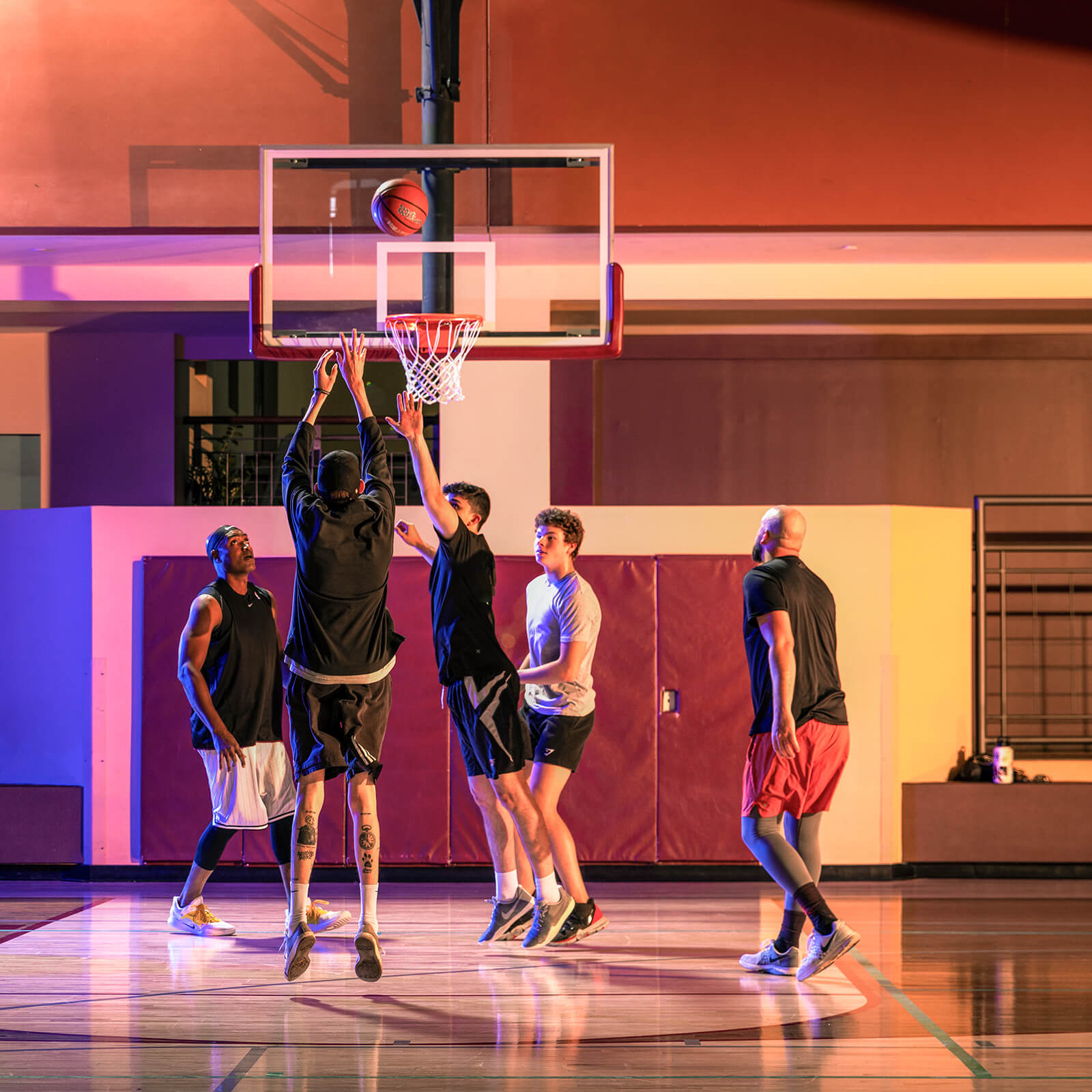 Village Clubs Camelback indoor basketball court with five men playing; two jump near the hoop as ball nears basket.