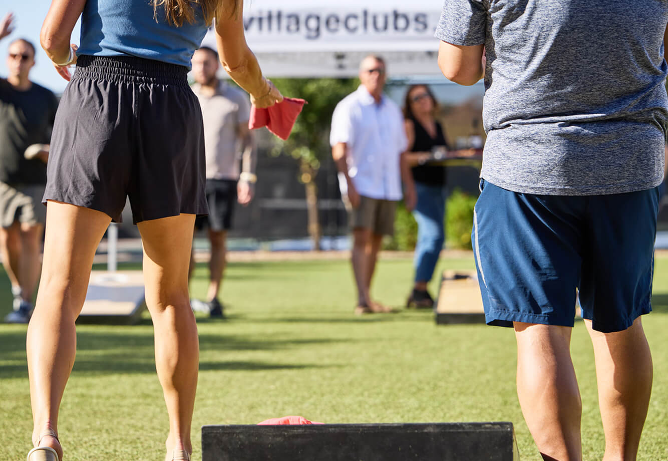 Members play cornhole at a sunny Village Clubs community event at DC Ranch, with others socializing near a branded banner.