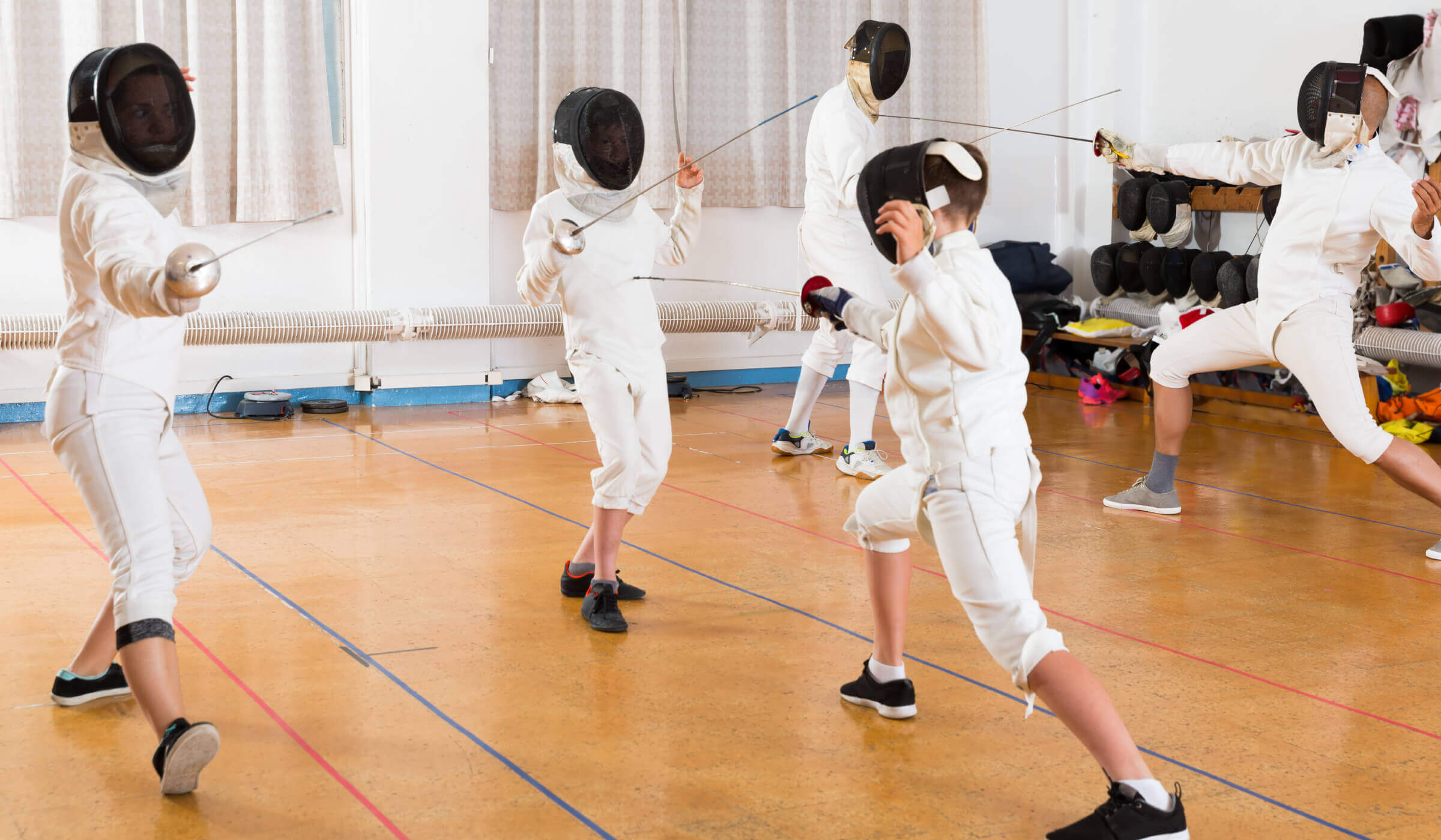 Children in fencing gear practice at Village Clubs Camelback gym, facing off with swords; equipment lines the wall in the background.