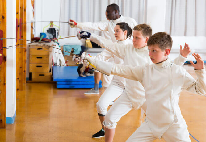 Members of Village Clubs at Camelback, including kids and adults, practice fencing lunges with foils in the gym.
