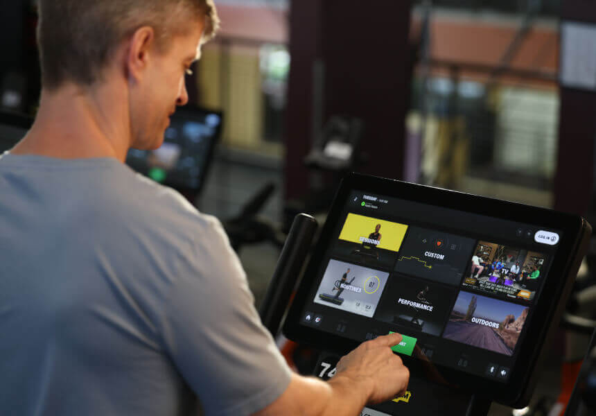 At Village Clubs Camelback, a member selects fitness programs on a touchscreen gym machine with modern equipment in the background.