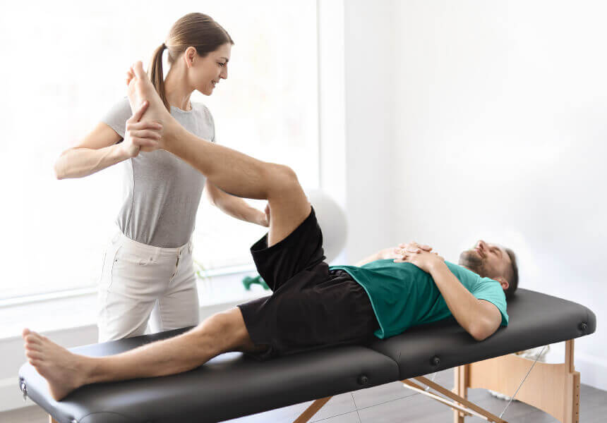A Village Clubs physical therapist at Camelback helps a man stretch his leg on a bright, sunlit treatment table.