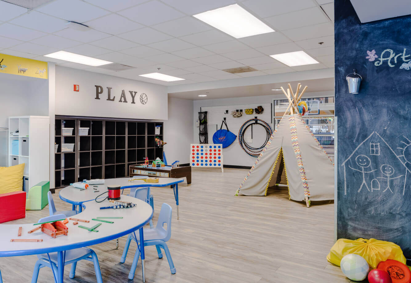 Village Clubs Camelback playroom with blue chairs, games, craft supplies, tipi tent, hula hoops, and a chalkboard with drawings.
