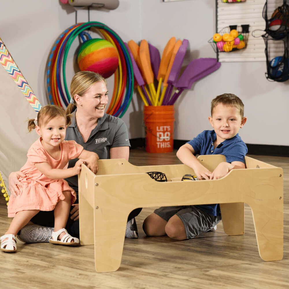 Children and an adult play with a wooden activity table at Village Clubs Camelback, surrounded by colorful indoor play equipment.