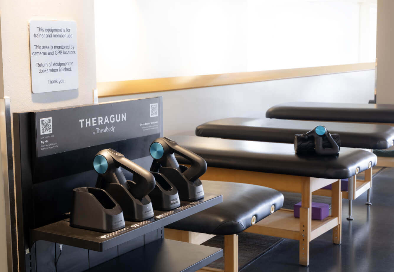 Village Clubs Camelback therapy room with tables and Theragun massagers; usage instructions displayed above the shelf.