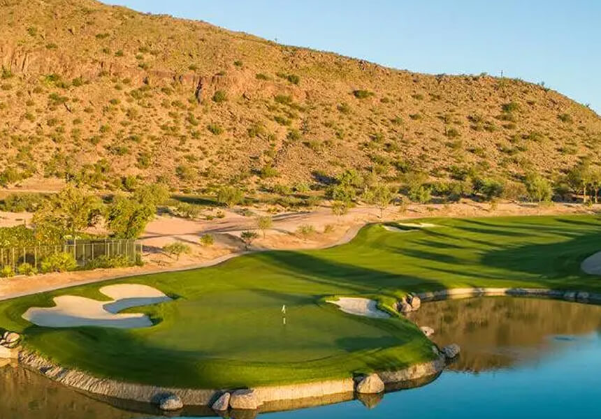 Village Clubs Camelback golf course with lush greens, sand bunkers, and pond set against Arizona desert hills under a blue sky.