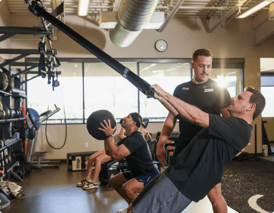 At Village Clubs Camelback, two men train with suspension straps and a medicine ball as a trainer guides them in a bright gym.