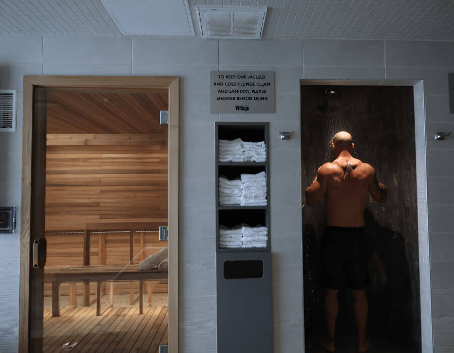 A man showers in a tiled stall beside a wooden sauna at Village Clubs Camelback, with white towels stacked neatly on nearby shelves.