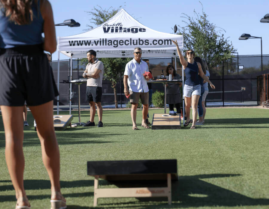 Guests play bean bag toss on the lawn near a Village Clubs tent at Camelback, with others socializing under a sunny Arizona sky.