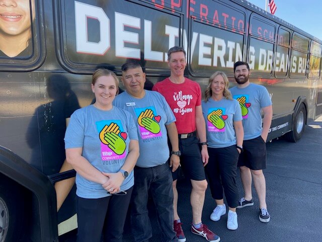 Five smiling people wearing Village Cares T-shirts pose by a “Delivering Dreams” bus at Village Clubs, Camelback location in Arizona.