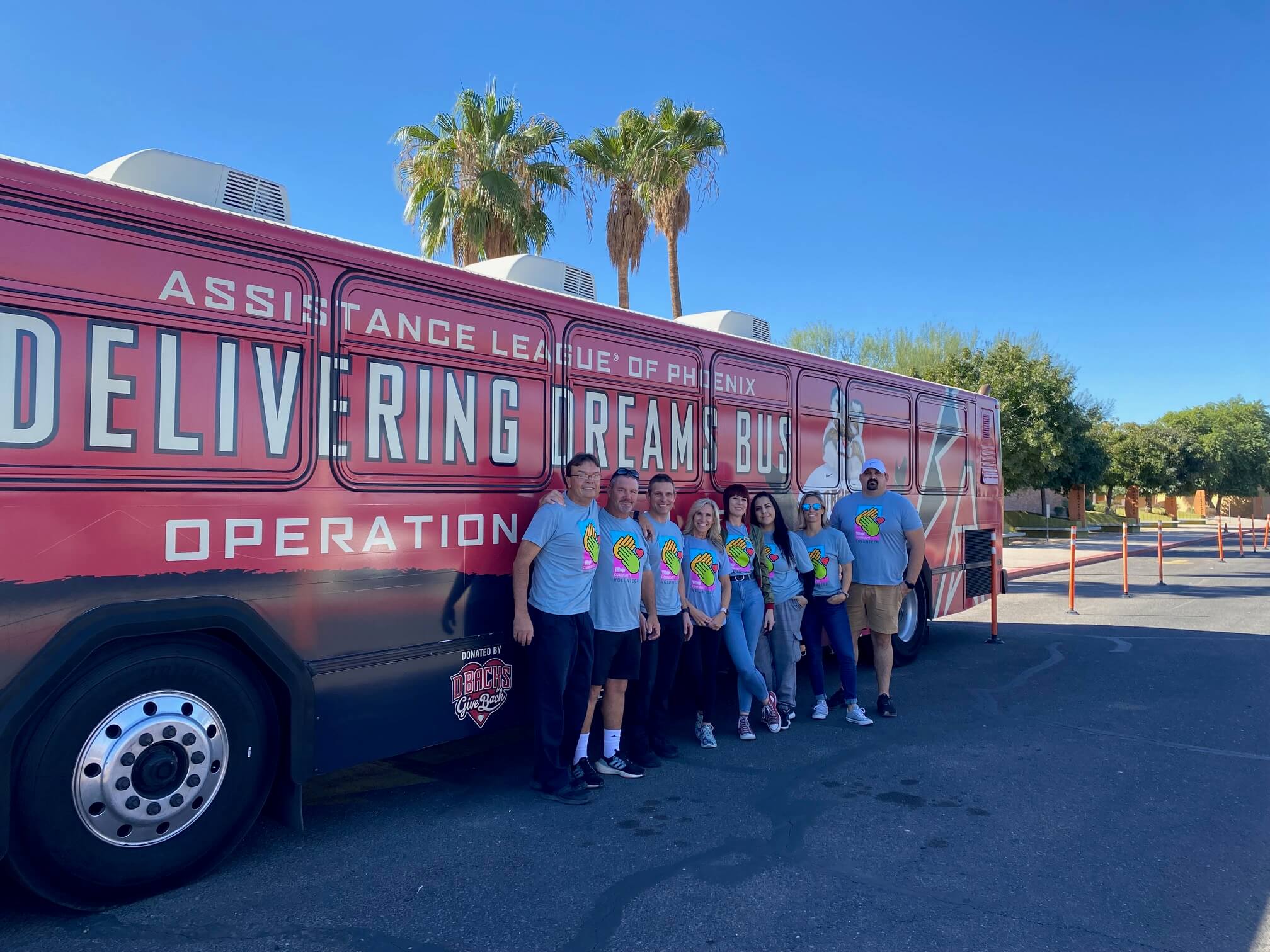 Village Clubs Camelback members in blue shirts gather by the Delivering Dreams Bus, supporting Phoenix community under palm trees.