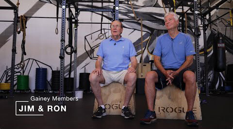 Two men in blue shirts sit on Rogue boxes in a Village Clubs Camelback gym, surrounded by fitness equipment. Members Jim & Ron.