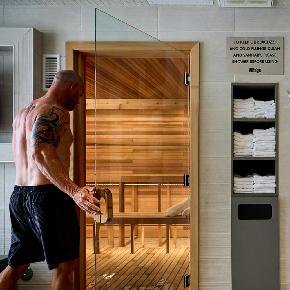 A muscular man opens a wooden sauna door at Village Clubs Camelback; white towels and membership info are neatly displayed nearby.
