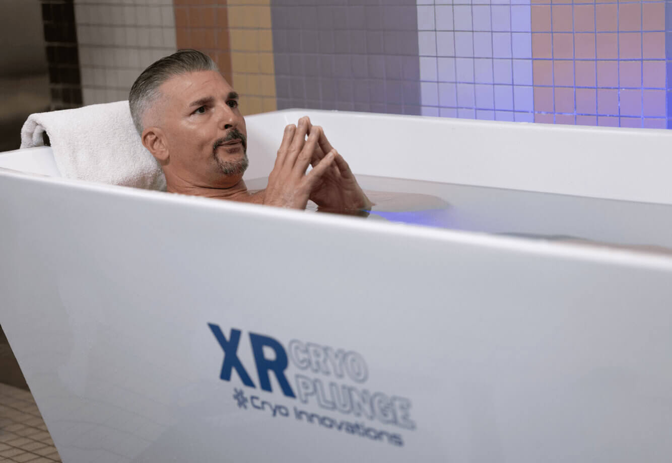 A man relaxes in a white XR Cryo Plunge tub at Village Clubs Camelback, enjoying membership benefits amid tiled walls.