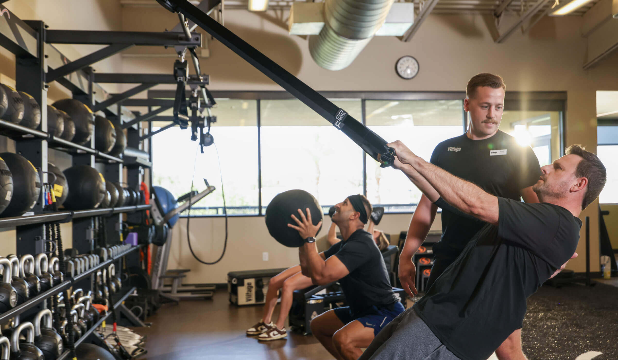 At Village Clubs Camelback, three men train using suspension straps and a medicine ball, with gym equipment in the background.