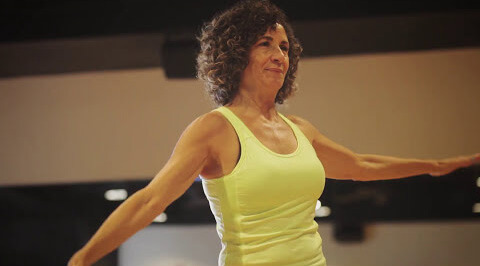 Courtney, in a yellow tank top, stretches her arms wide at Village Clubs Camelback, Arizona, smiling in a bright fitness studio.