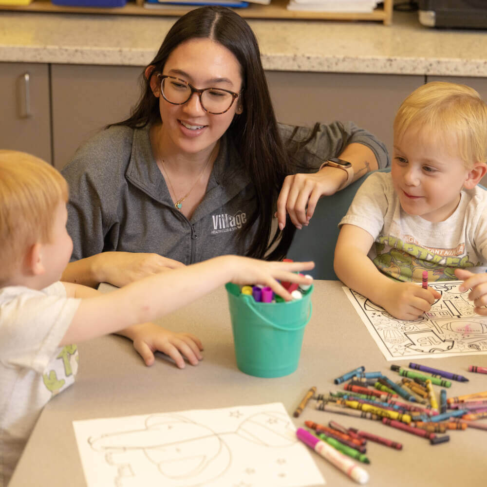 At Village Clubs Camelback, a woman and two children smile as they color together with crayons and markers at a craft table.
