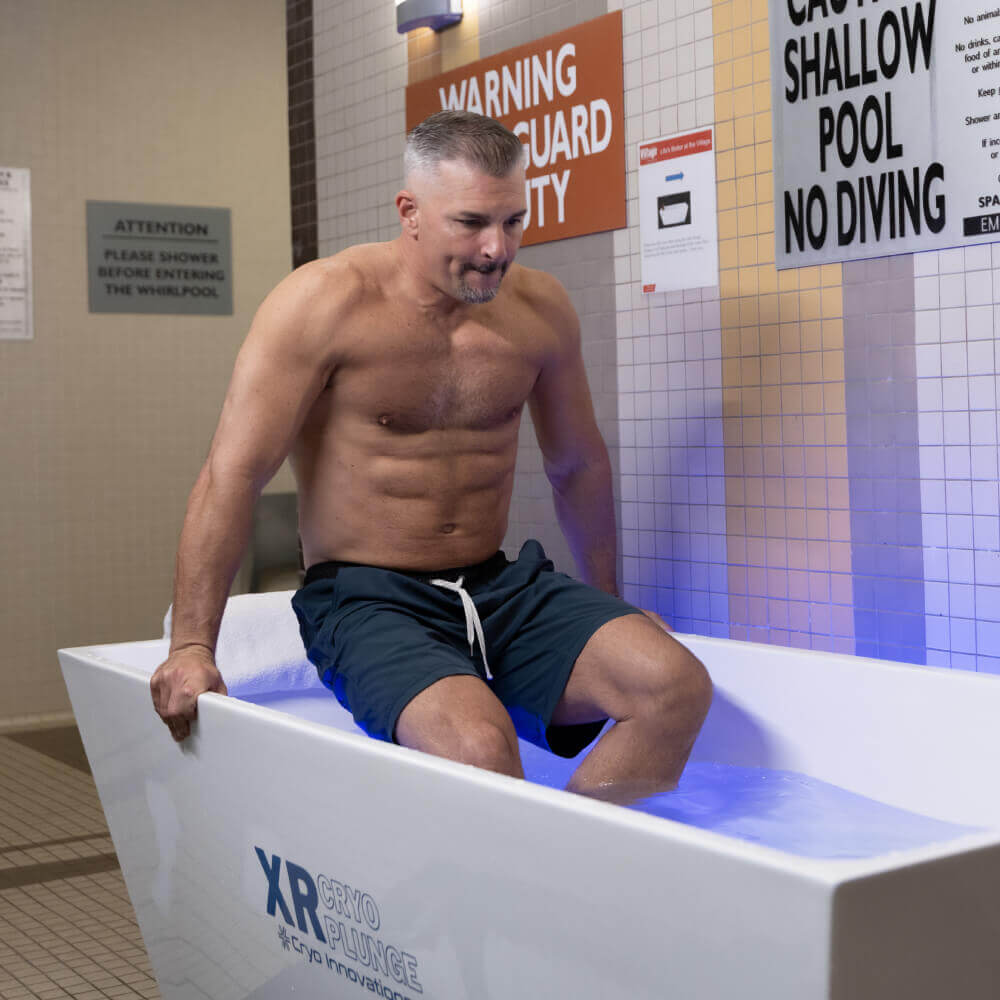 A fit man prepares to use the cold plunge tub at Village Clubs Camelback's indoor pool, with tiled walls and safety signs visible.