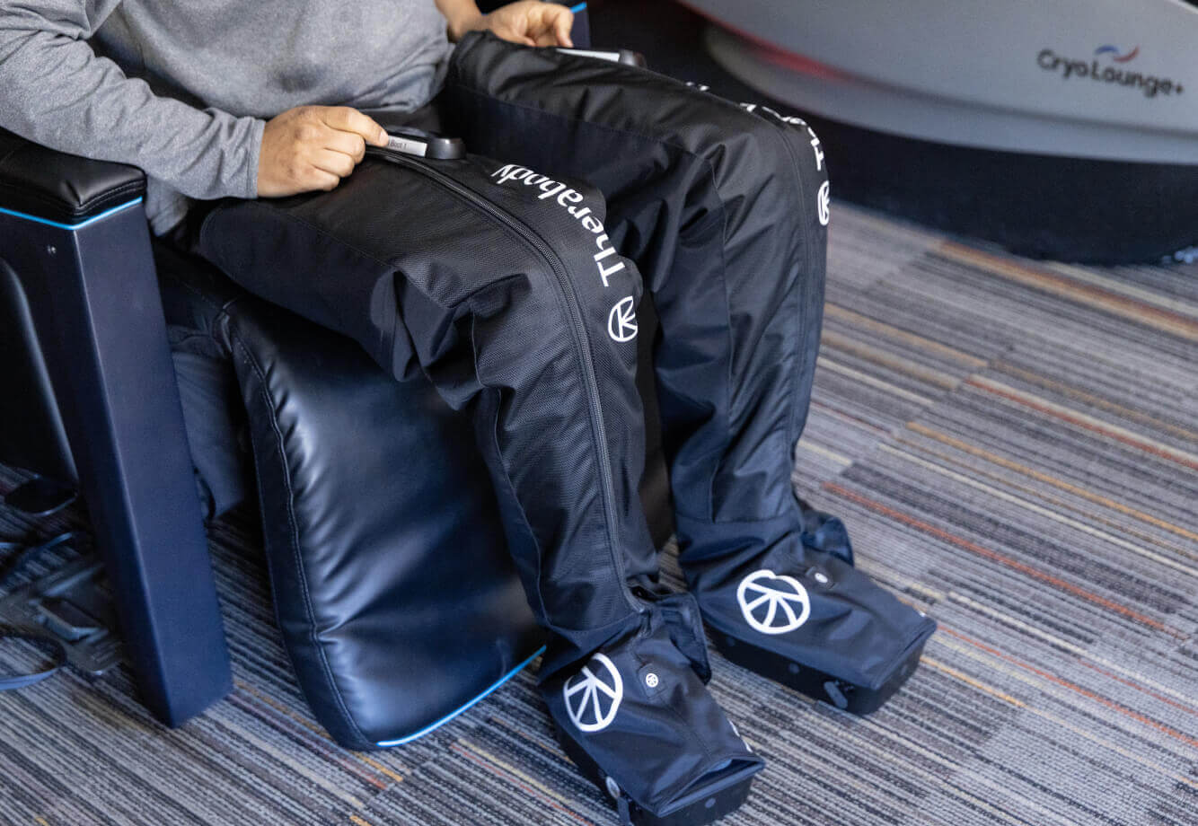 At Village Clubs Camelback, a person uses Therabody compression leg sleeves while seated on a striped carpeted floor.