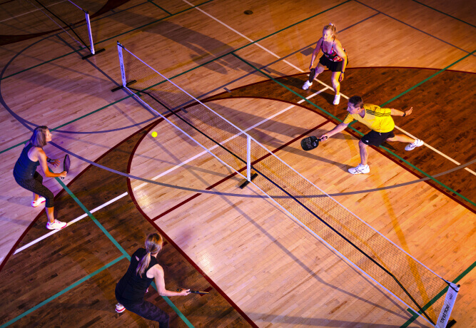 Four players enjoy doubles pickleball at Village Clubs Camelback, Arizona, on a lined indoor court with paddles and net.