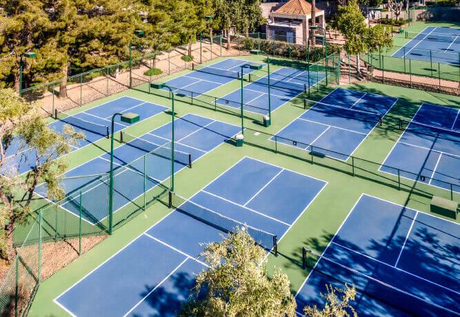 Aerial view of Village Clubs pickleball courts at Camelback, featuring blue and green surfaces, fencing, and nearby trees.