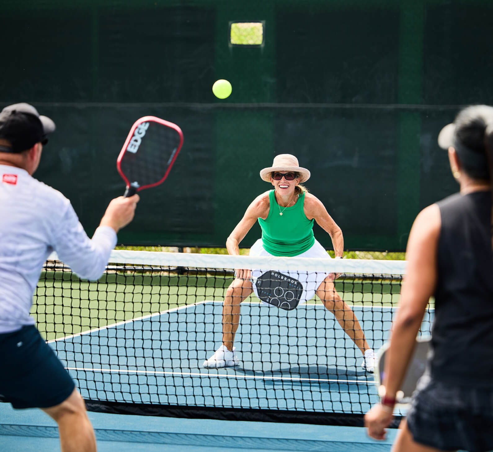 Members enjoy a pickleball match at Village Clubs Camelback on a blue court, as a woman in green returns the ball over the net.