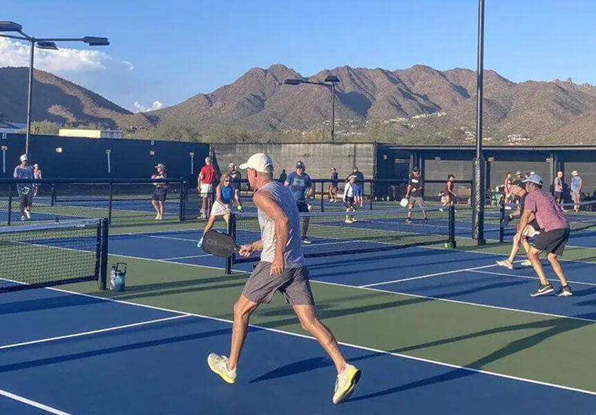 Members enjoy pickleball on sunny outdoor courts at Village Clubs Camelback, with mountain views and players in athletic gear.