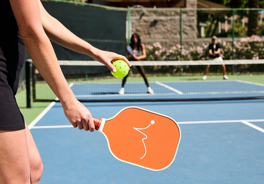 Close-up of a player at Village Clubs Camelback holding an orange paddle and yellow ball, with two ready players across the pickleball net.
