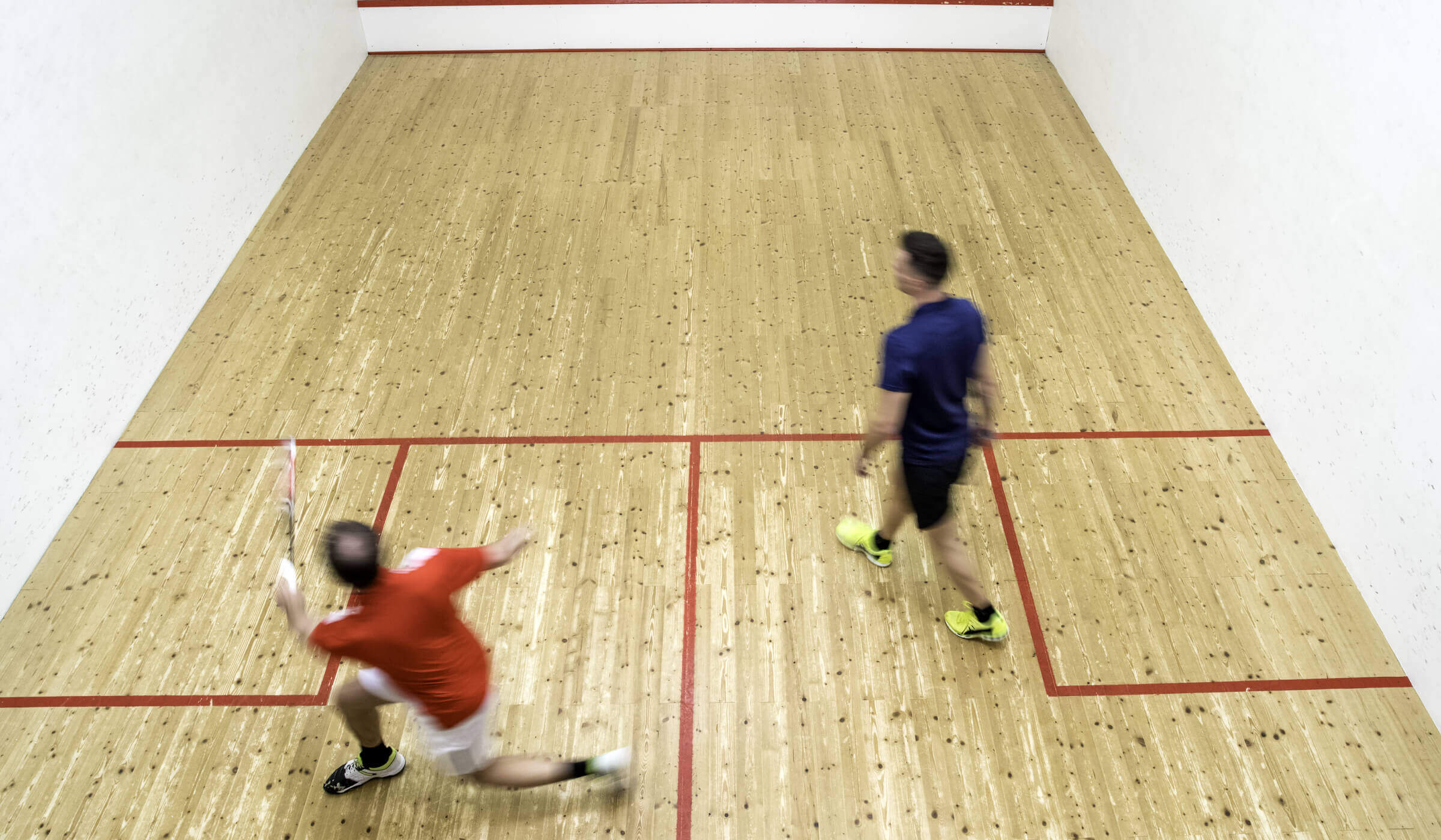 Two men play squash at Village Clubs Camelback, one lunging to hit the ball as the other watches, both in athletic wear on court.