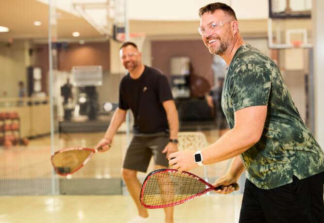 Two men play racquetball at Village Clubs Camelback in Arizona, wearing safety glasses and sporty attire on the indoor court.