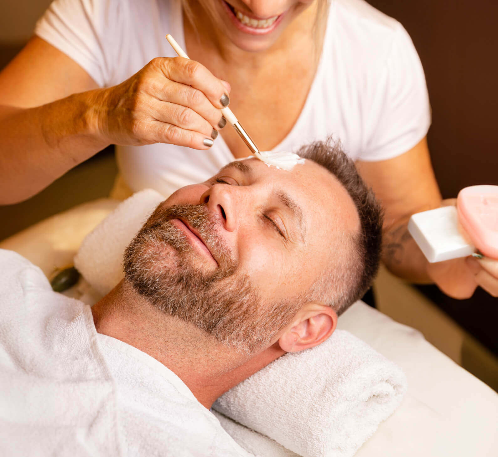 A bearded man enjoys a facial mask at Village Clubs Camelback spa, with a professional gently applying the treatment.