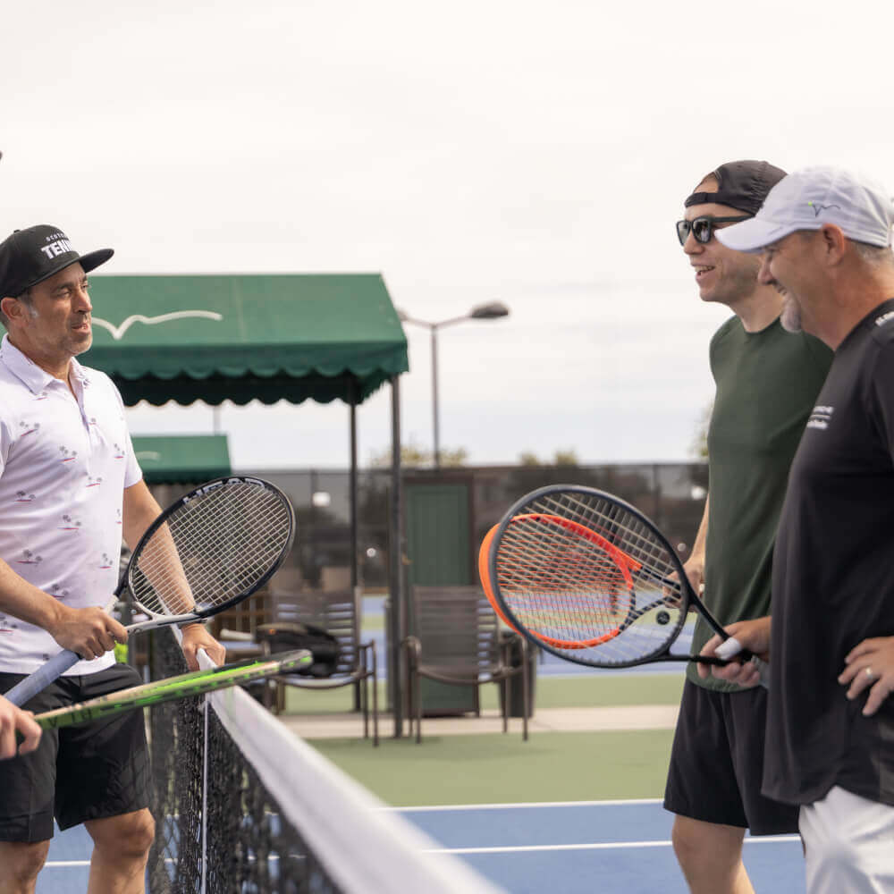 Four men enjoy a friendly chat by the net on a Village Clubs tennis court at Camelback, Arizona, holding racquets on a sunny day.