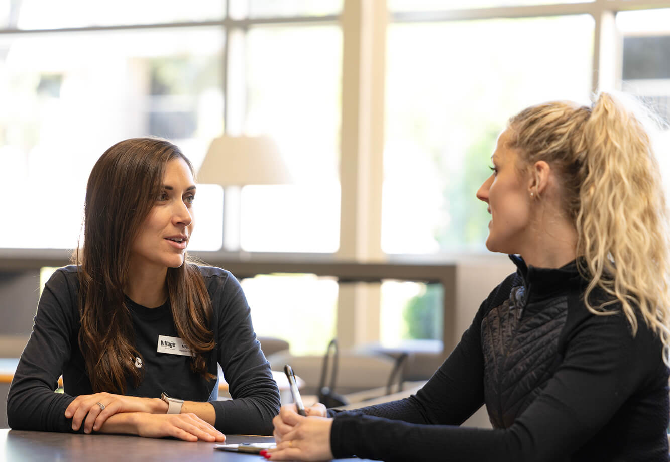Two women engage in conversation at Village Clubs Camelback, seated indoors with natural light streaming through large windows.