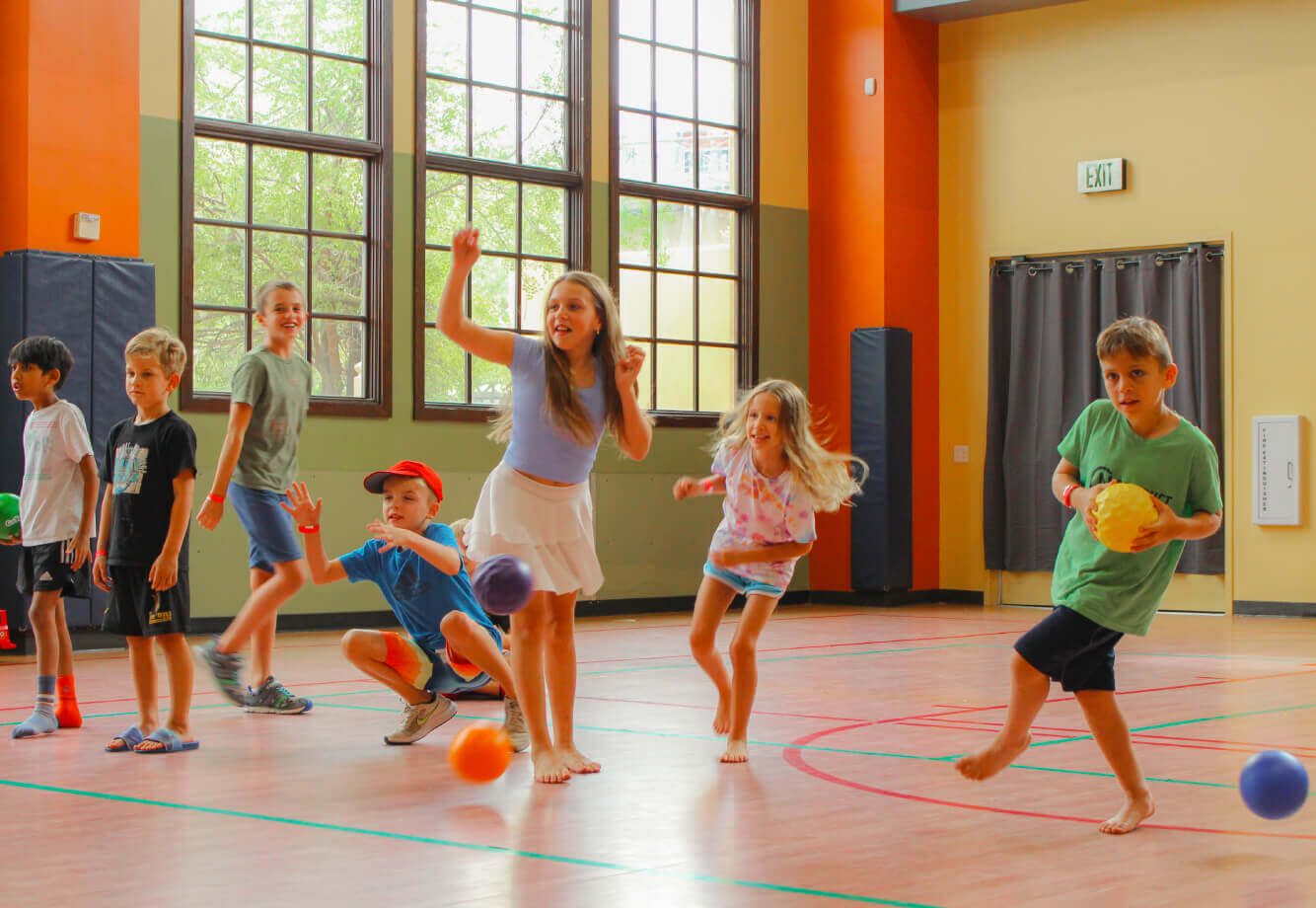 Kids play dodgeball in a sunlit Village Clubs gym at Camelback, enjoying Youth Programs with energy and smiles.