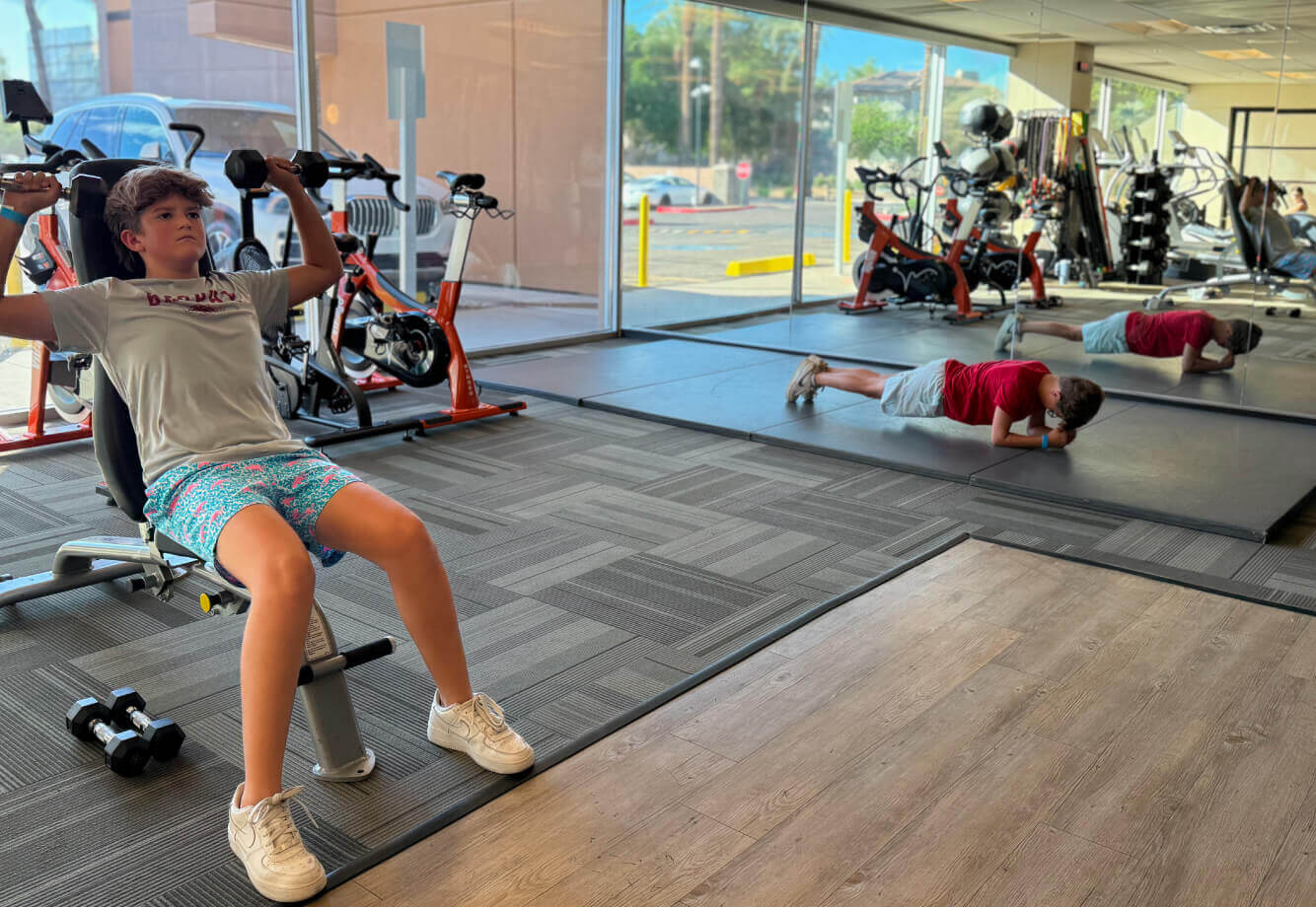 At Village Clubs Camelback, two boys join youth fitness: one uses a shoulder press, another in red holds a plank in the gym.
