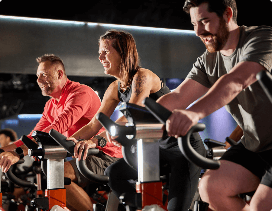 Three adults enjoy a group cycling class at Village Clubs Camelback, smiling as they ride stationary bikes in the gym.
