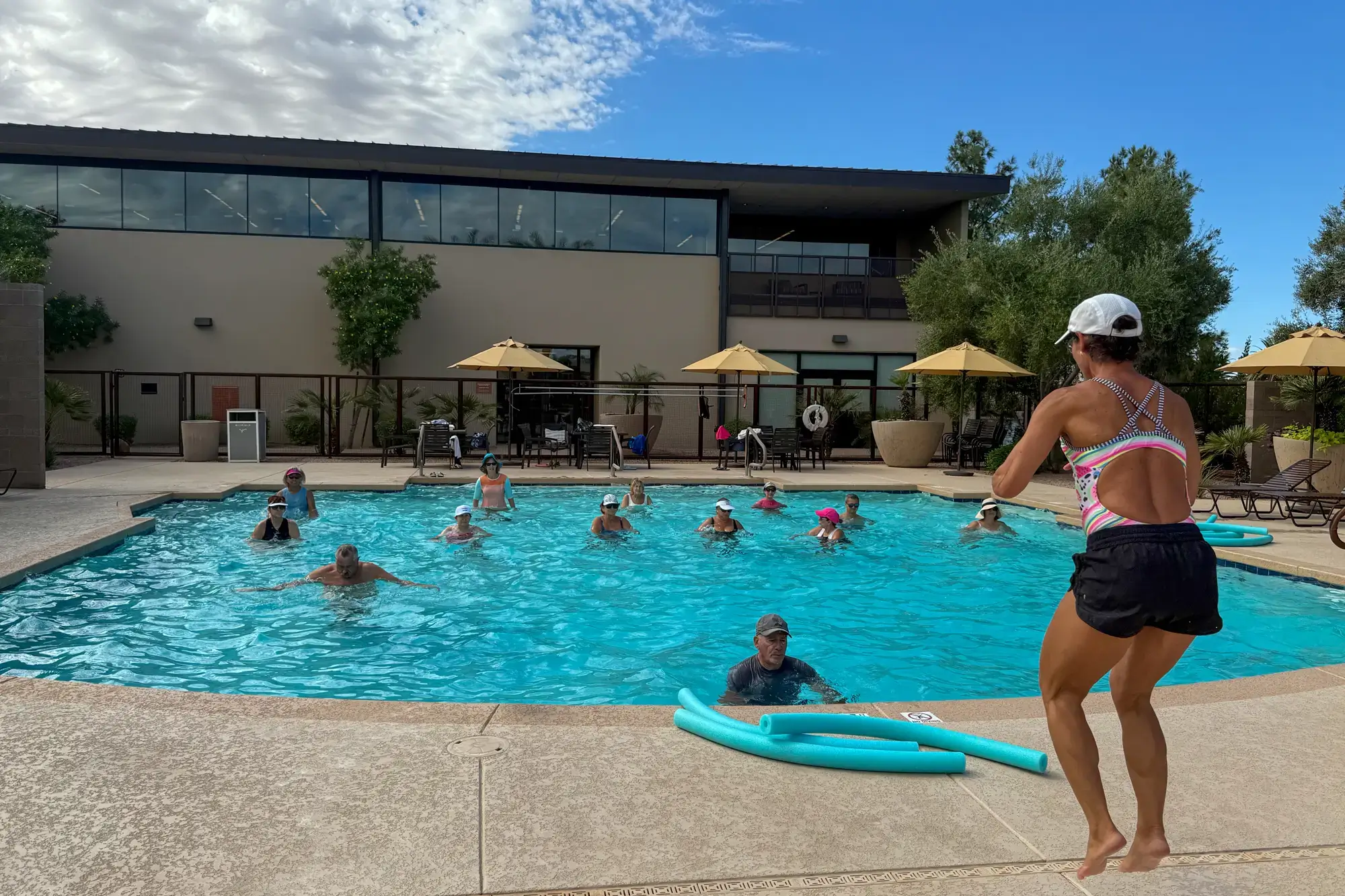 A Village Clubs instructor leads a water aerobics class at the Camelback outdoor pool with adults and poolside lounge chairs nearby.