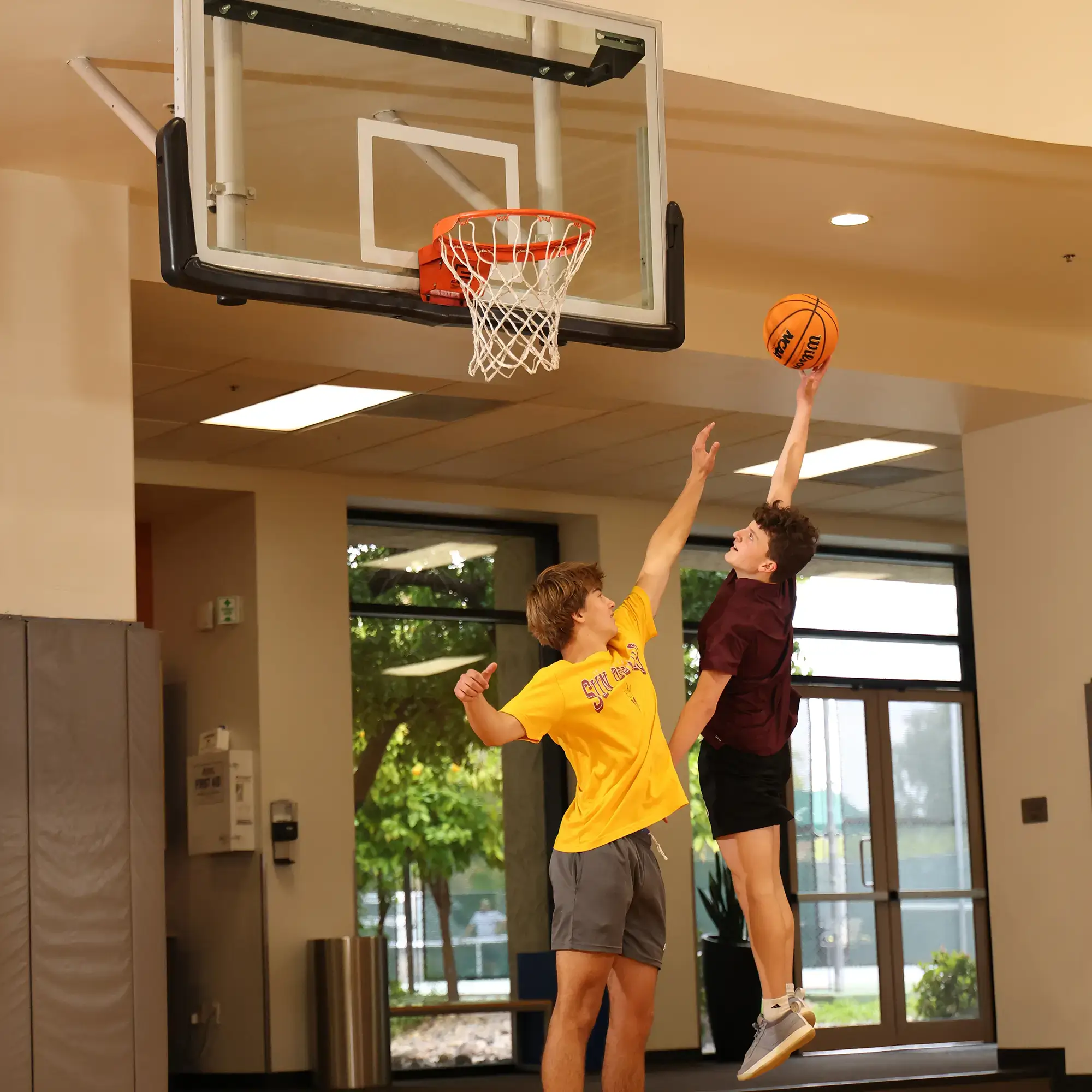 Teens play basketball in Village Clubs’ indoor court at Camelback, shooting and defending with trees visible through large windows.